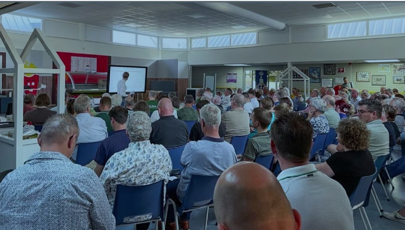 Groepsbijeenkomst met presentatie over complexe opgaven en belangen, scherm toont onleesbare tekst, vlaggen en borden in zaal Deventer